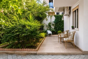 A patio with a table and chairs, surrounded by lush greenery and a white wall. - Bubu Garden Apartament - 3 Bedrooms (Durres)