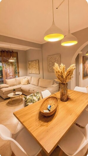 A modern living room with a wooden dining table, white chairs, a beige sofa, and a large window with curtains. - Bubu Garden Apartament - 3 Bedrooms (Durres)