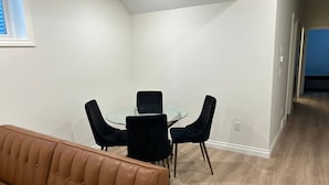 A room with a glass-top table, four chairs, a brown sofa, and a wooden floor. - Two Bed Room & One Bathroom Secondary Suit (Calgary)