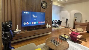 Een moderne woonkamer met een flatscreen televisie, een klok, een bank, een salontafel en een eethoek op de achtergrond.