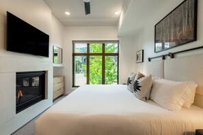 Ein modernes Schlafzimmer mit einem großen Bett, einem Kamin, einem Fernseher und einem großen Fenster mit Blick auf Grünflächen.