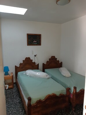 Ein kleines Zimmer mit zwei Holzbett, einem Nachttisch mit Lampe, einem gerahmten Bild an der Wand und einer Deckenleuchte.