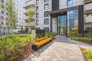 A modern building with a paved walkway, a wooden bench, and green plants. - Spacious 3-Bedroom Apartment | Air Conditioning | Garage | Terrace (Warszawa)