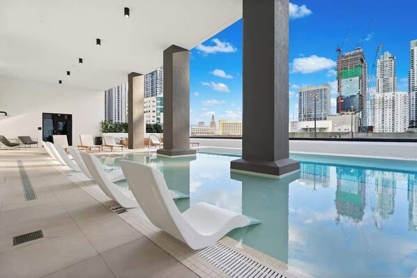 Ein Rooftop-Infinity-Pool mit modernen weißen Stühlen, großen Säulen und Blick auf eine Stadtlandschaft.