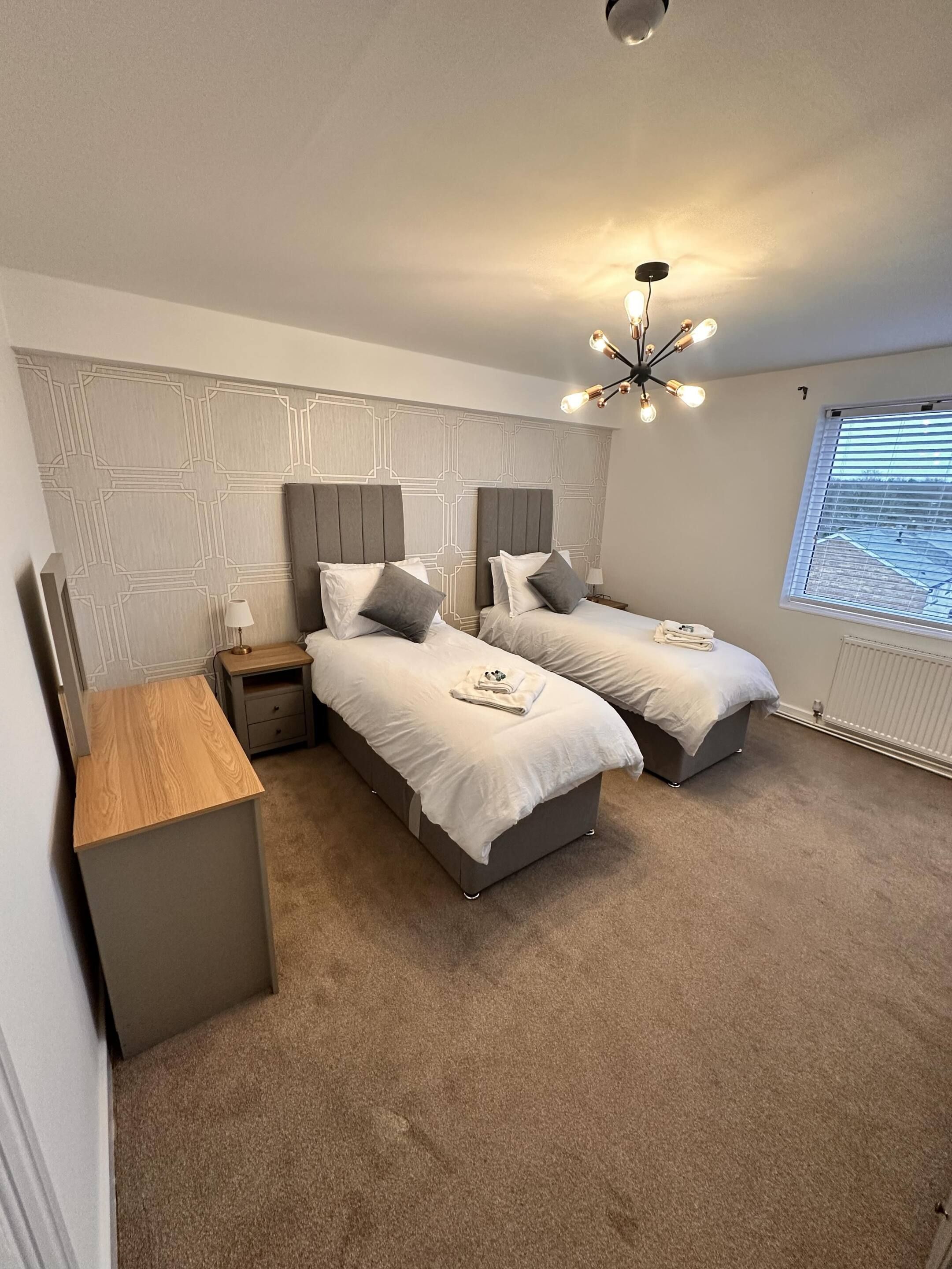 A bedroom with two beds, a bedside table, a lamp, a ceiling light fixture, and a window with blinds.