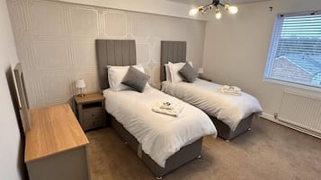 A bedroom with two beds, a bedside table, a lamp, a ceiling light fixture, and a window with blinds.