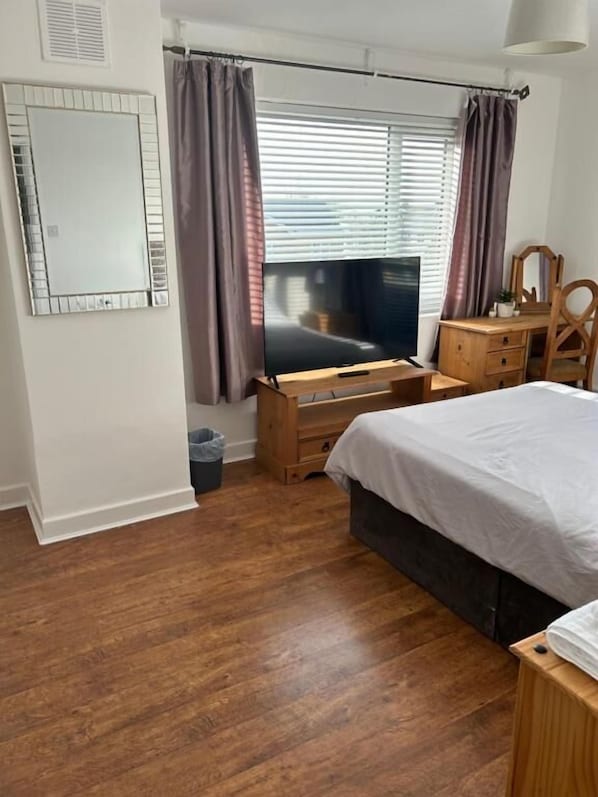 Un dormitorio con cama, televisor, cómoda de madera, espejo y ventana con cortinas.