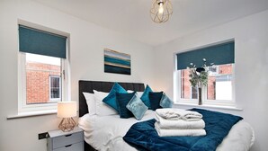 A bedroom with a bed, nightstand, lamp, and windows with views of a brick building. - Captains Retreat, Whitby (Whitby)