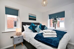 A bedroom with a bed, nightstand, lamp, and windows with views of a brick building. - Captains Retreat, Whitby (Whitby)