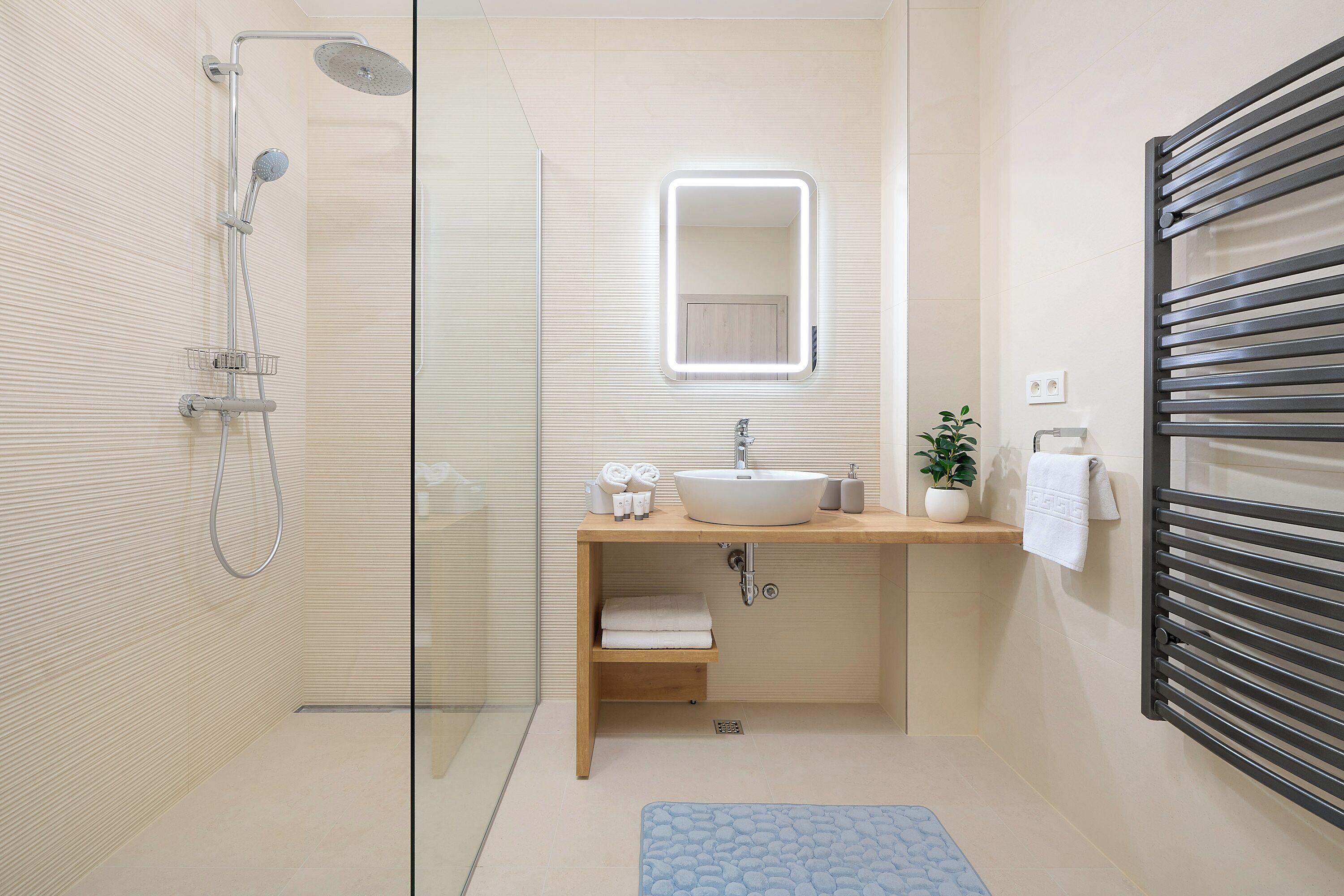 Un baño moderno con cabina de ducha de vidrio, un lavabo blanco, un espejo, un perchero para toallas y una planta pequeña.