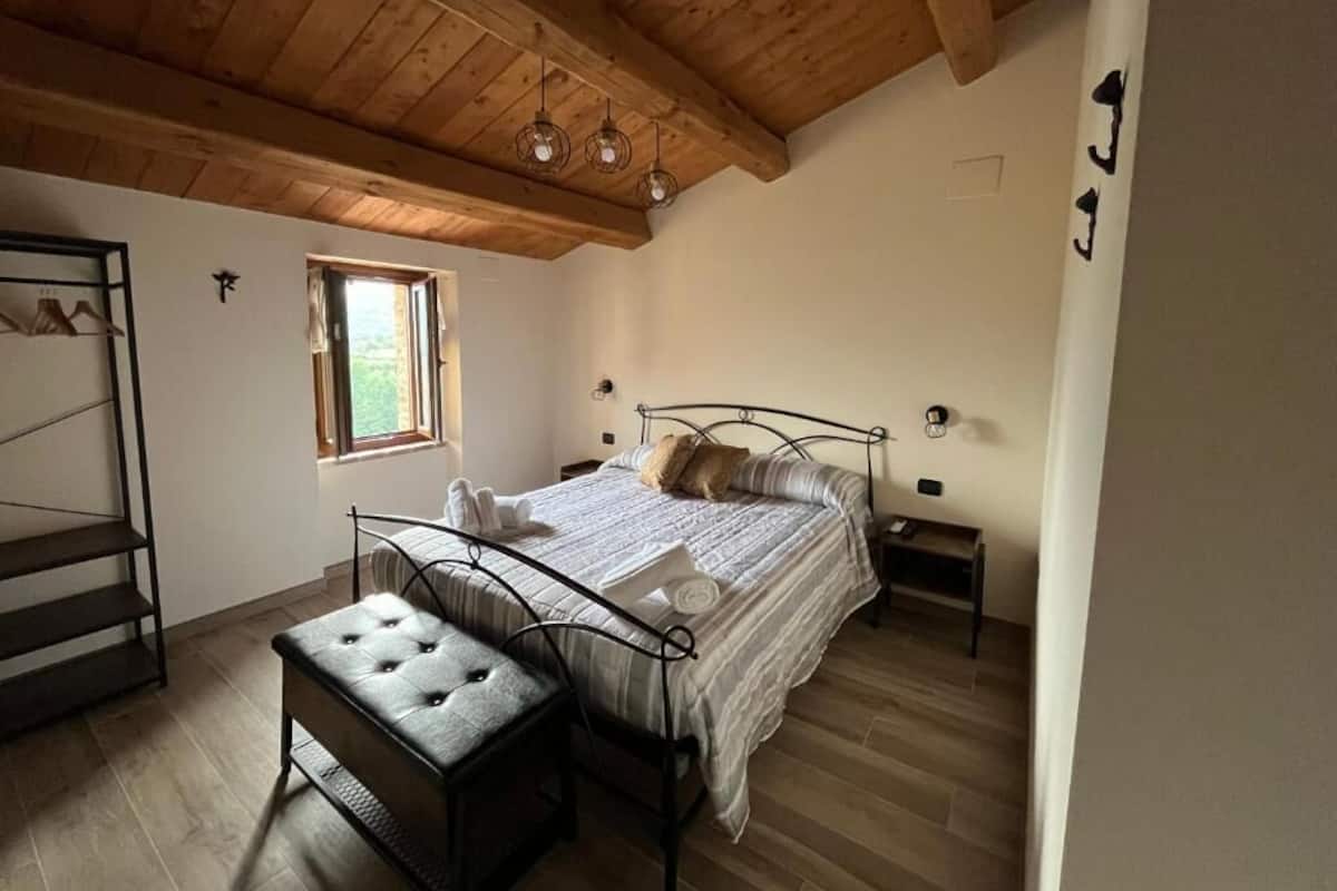 A bedroom with a bed, a bench, a window, and a wooden ceiling.
