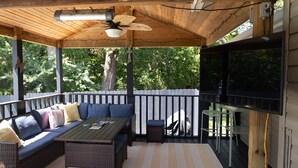 Eine überdachte Terrasse mit einer Couch, einem Tisch und Stühlen, einem Deckenventilator und einem Fernseher.