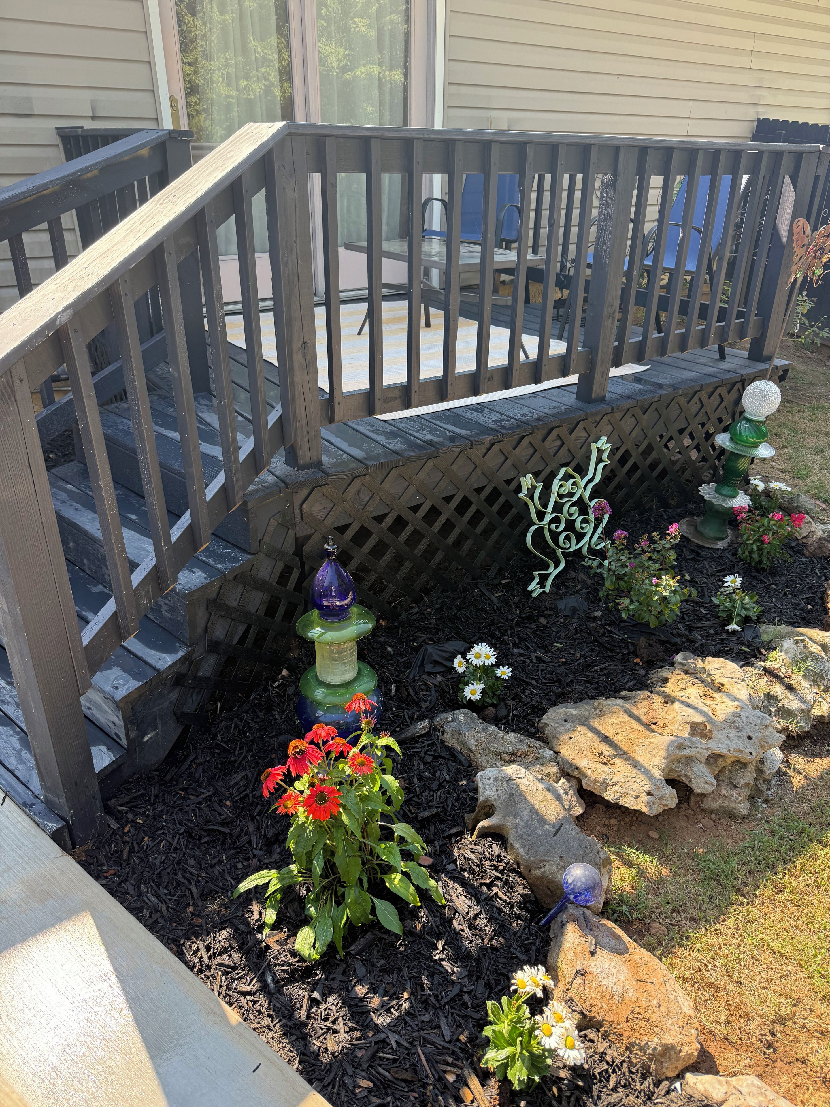 Une terrasse en bois avec une rambarde, un jardin fleuri et un abreuvoir pour les oiseaux, et une maison en arrière-plan.