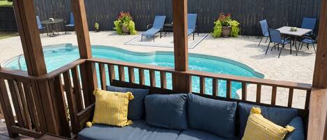 A covered outdoor seating area with a pool, wooden furniture, and a view of the backyard.