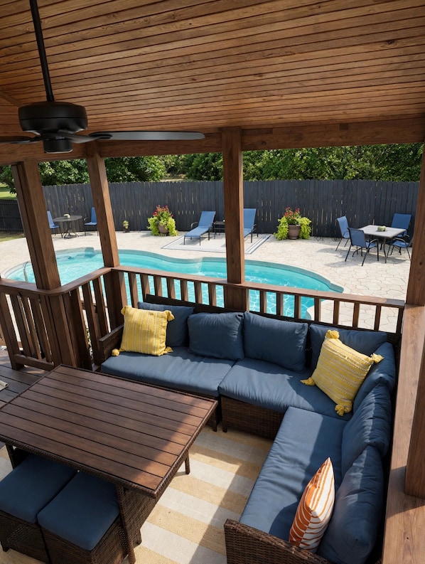 Un espacio de estar al aire libre cubierto, con piscina, muebles de madera y vista al jardín trasero.