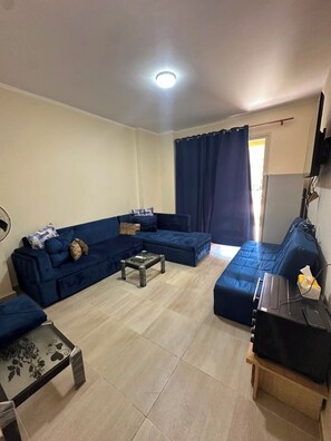A living room with a sectional sofa, a coffee table, a TV on a stand, and a curtain. -   شاليه عائلي فاخر  بجاردن في AquaView – أكوافيو (elhammam)