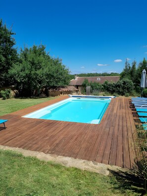 En rektangulær swimmingpool med en træterrasse, omgivet af græs og træer.