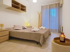 Ein ordentlich eingerichtetes Schlafzimmer mit Bett, Nachttischen, einer Lampe, einem Fenster mit Vorhängen und einer Kerze auf einem Tablett.
