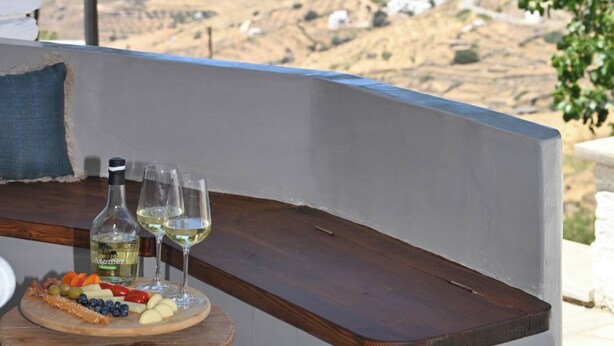 A table with a bottle of wine and glasses, a wooden board with snacks, and a scenic view of the ocean and hills. - Katoi Suite, Vivian Suites Tinos (Tinos)