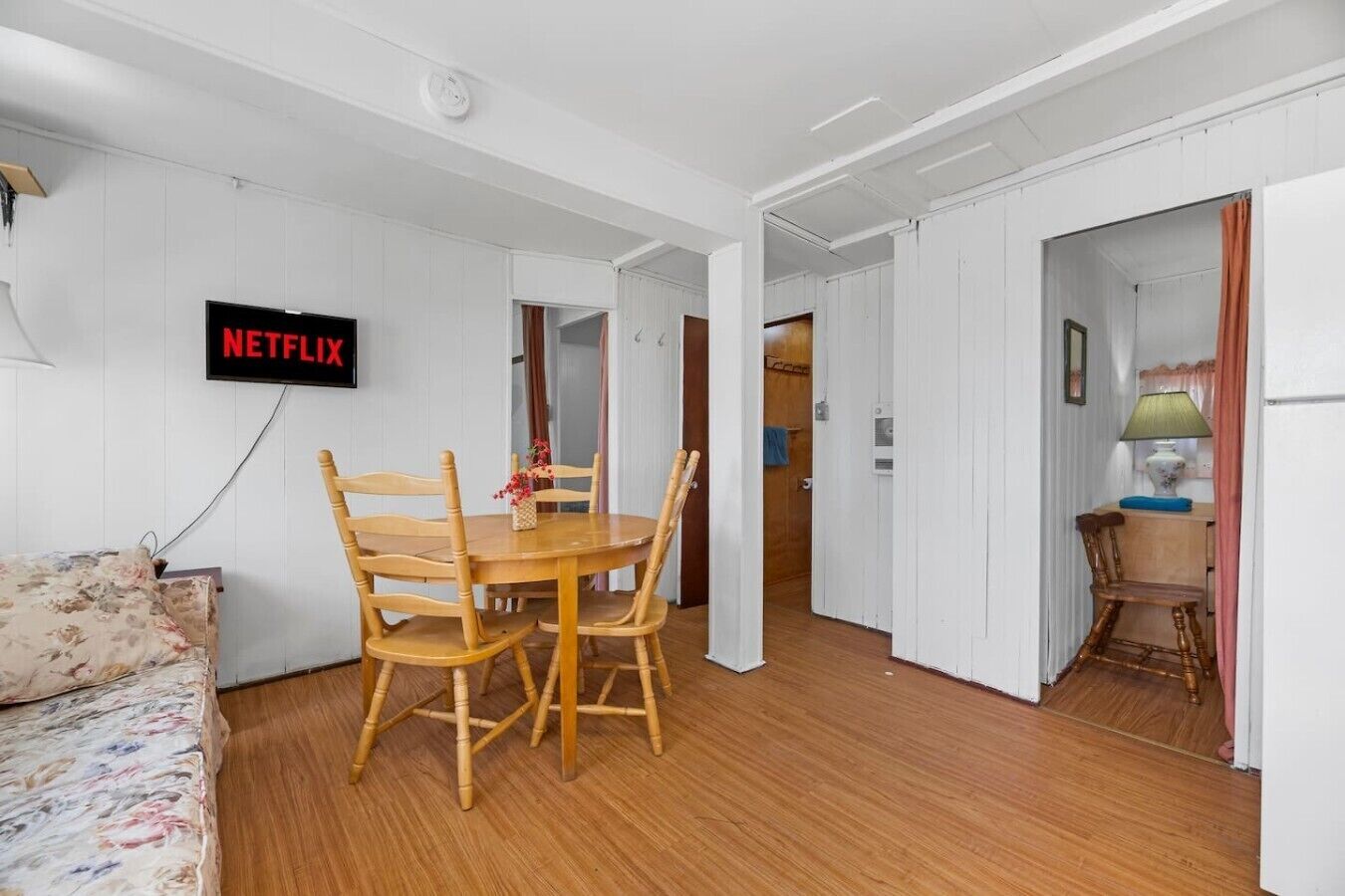 Habitació amb taula de dinar de fusta, cadirees, un llit amb roba de llenceria floral i un televisor fixat a la paret que mostra 'NETFLIX', tot amb parets de panells blancs.