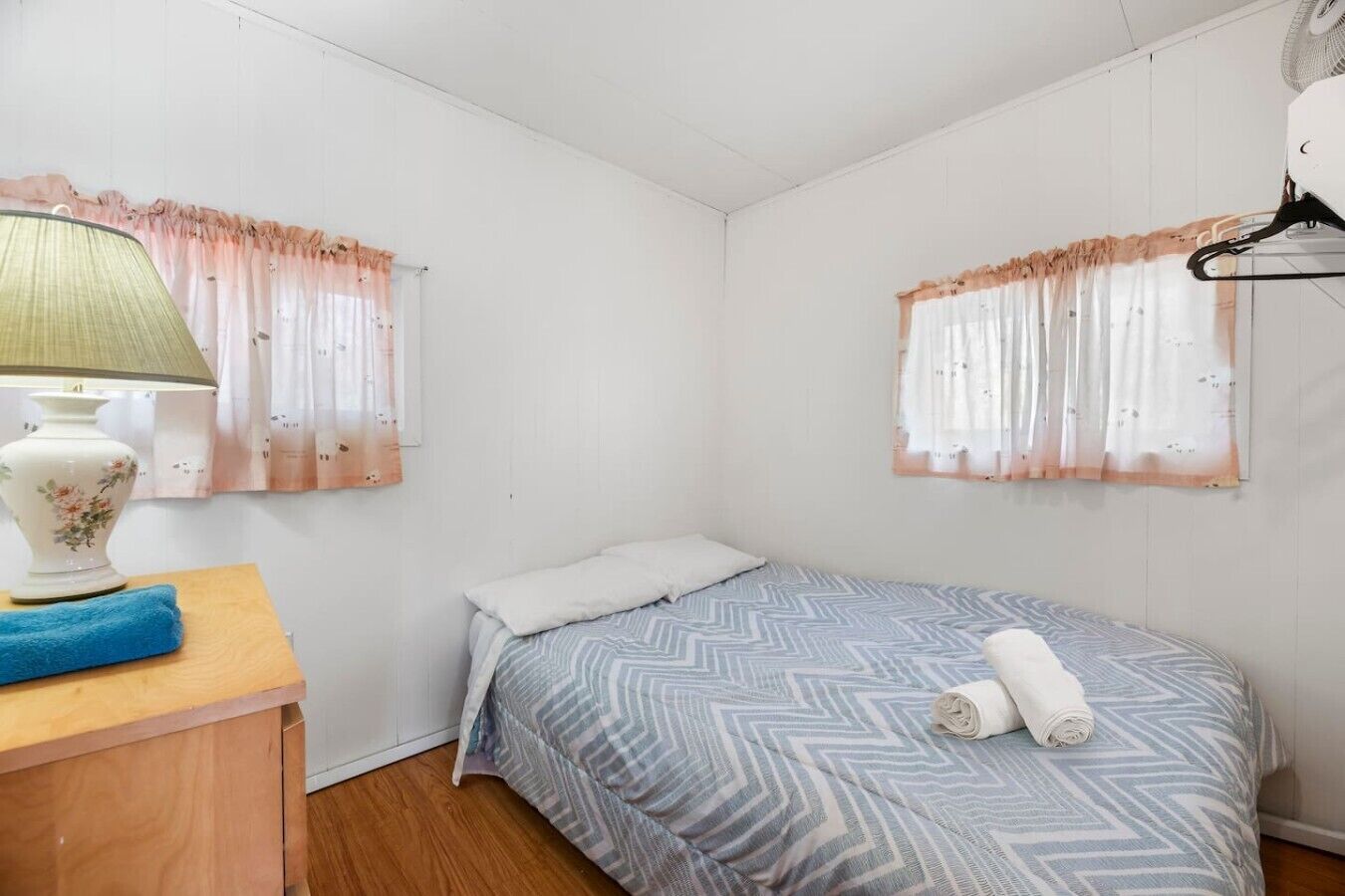 A small, single-bed room with a wooden nightstand, a lamp, and a window with sheer curtains.