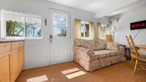 Ein Zimmer mit einer weißen Tür, einem blumigen Sofa, einem Holztisch, einem Stuhl und einem an der Wand montierten Fernseher, der 'NETFLIX' anzeigt.