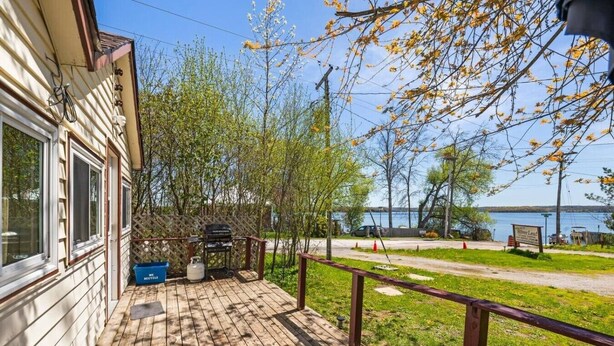 Eine Holzterrasse mit Grill, einem blauen Behälter mit der Aufschrift 'WELLE RESTAUBE' und Blick auf einen See mit Bootssteg.