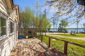 Una terraza de madera con una parrilla, un contenedor azul que dice 'WELLE RESTAUBE' y vista a un lago con un muelle para botes.