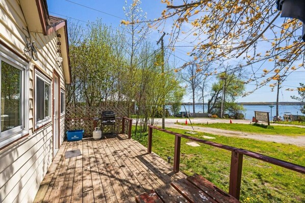 Una terraza de madera con una parrilla, un contenedor azul que dice 'WELLE RESTAUBE' y vista a un lago con un muelle para botes.
