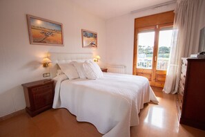 Ein Schlafzimmer mit einem Bett, zwei Bildern, einem Nachttisch, einer Kommode und Blick durch ein Fenster.