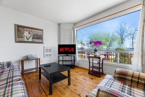 A living room with a black coffee table, plaid sofas, a TV displaying 'NETFLIX,' and a balcony with a view of trees and a fence.
