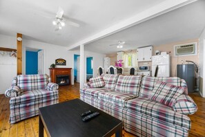 A living room with a plaid sofa, a fireplace, a ceiling fan, and a kitchen area with cabinets and appliances.