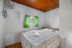 A single bed with a white pillow and a rolled towel, a window with green curtains, a wall-mounted fan, and a power outlet.