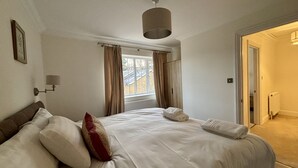 Un dormitori amb llit, finestra amb cortines, làmpada penjada a la paret i porta que dona a una altra habitació.