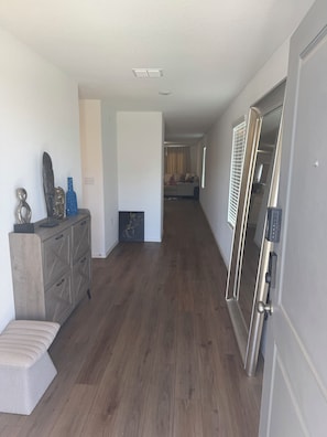 A hallway with a wooden floor, a grey door with a keypad lock, and a small cabinet with decorative items. - Spacious Modern Home w/ 98” TV + Fast WiFi (Pflugerville)