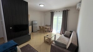 Ein Wohnzimmer mit einer Couch, einem Couchtisch, einem Fernseher, einem Kühlschrank, einem Esstisch mit Stühlen und einer Glasschiebetür, die auf einen Balkon führt.