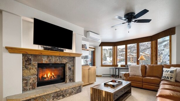 A living room with a stone fireplace, a flat-screen TV, a ceiling fan, a brown leather sofa, and a view of a snowy landscape through large windows. - Stylish ski-in/out condo w/views in base area next to gondola, pool & restaurant (Steamboat Springs)