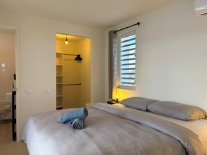 Ein Schlafzimmer mit Bett, Nachttisch, Fenster mit Jalousien, Kleiderschrank mit Regalen und einer Tür zu einem anderen Raum.