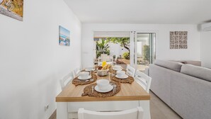 Uma sala de jantar com uma mesa de madeira posta para quatro pessoas, cadeiras brancas e um sofá cinzento ao fundo.