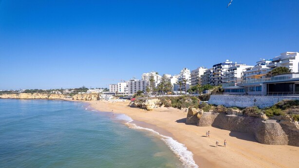 Uma praia costeira com edifícios, céu claro e areal arenoso.