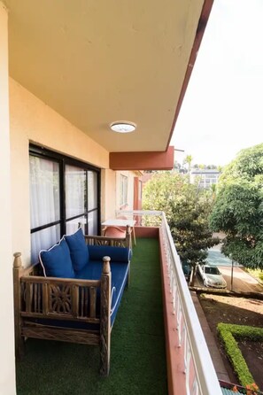 A balcony with a wooden sofa, a small table, and a view of a garden and street. - Gorgeous 3-bedroom apartment with AC in vibrant Nairobi (Nairobi)