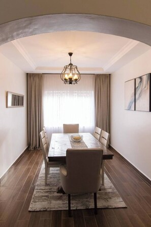 A dining room with a marble table, beige chairs, a chandelier, and a large window with curtains. - Gorgeous 3-bedroom apartment with AC in vibrant Nairobi (Nairobi)