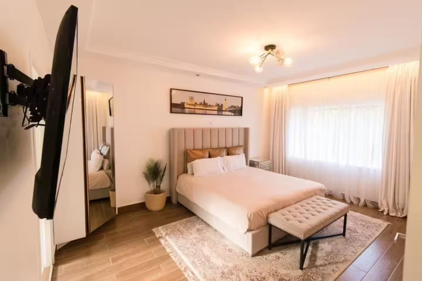 A bedroom with a large bed, a bedside table, a potted plant, a mirror, and a painting on the wall.