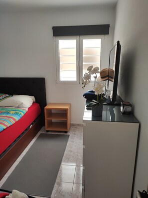 Een slaapkamer met een bed, een bureau, een stoel, een raam en een klein houten plankje.