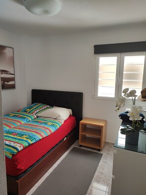Un dormitorio con cama, mesita de noche, ventana con persianas y estante en la pared.