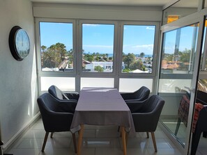 Een eethoek met een tafel en stoelen, grote ramen met uitzicht op zee en gebouwen, en een wandklok.