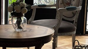 Ein Zimmer mit einem runden Holztisch, einem Stuhl mit floralem Muster und Blick durch ein Fenster mit einem Metallgitter.