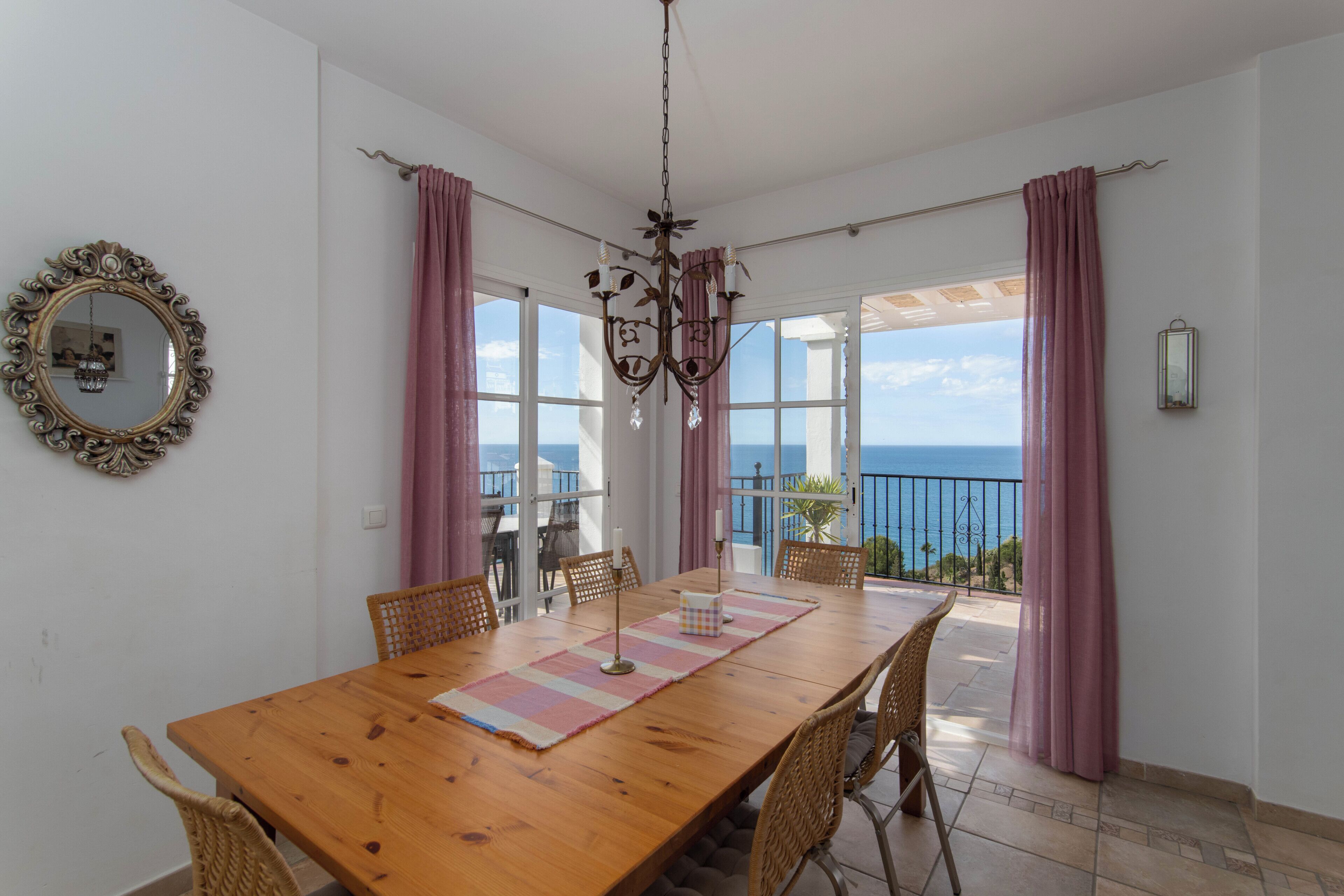 Un coin repas avec une table en bois, des chaises en osier, un lustre et une vue sur la mer grâce à de grandes fenêtres.