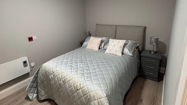 Un dormitorio con cama, mesitas de noche, enchufe en la pared y luz en el techo.