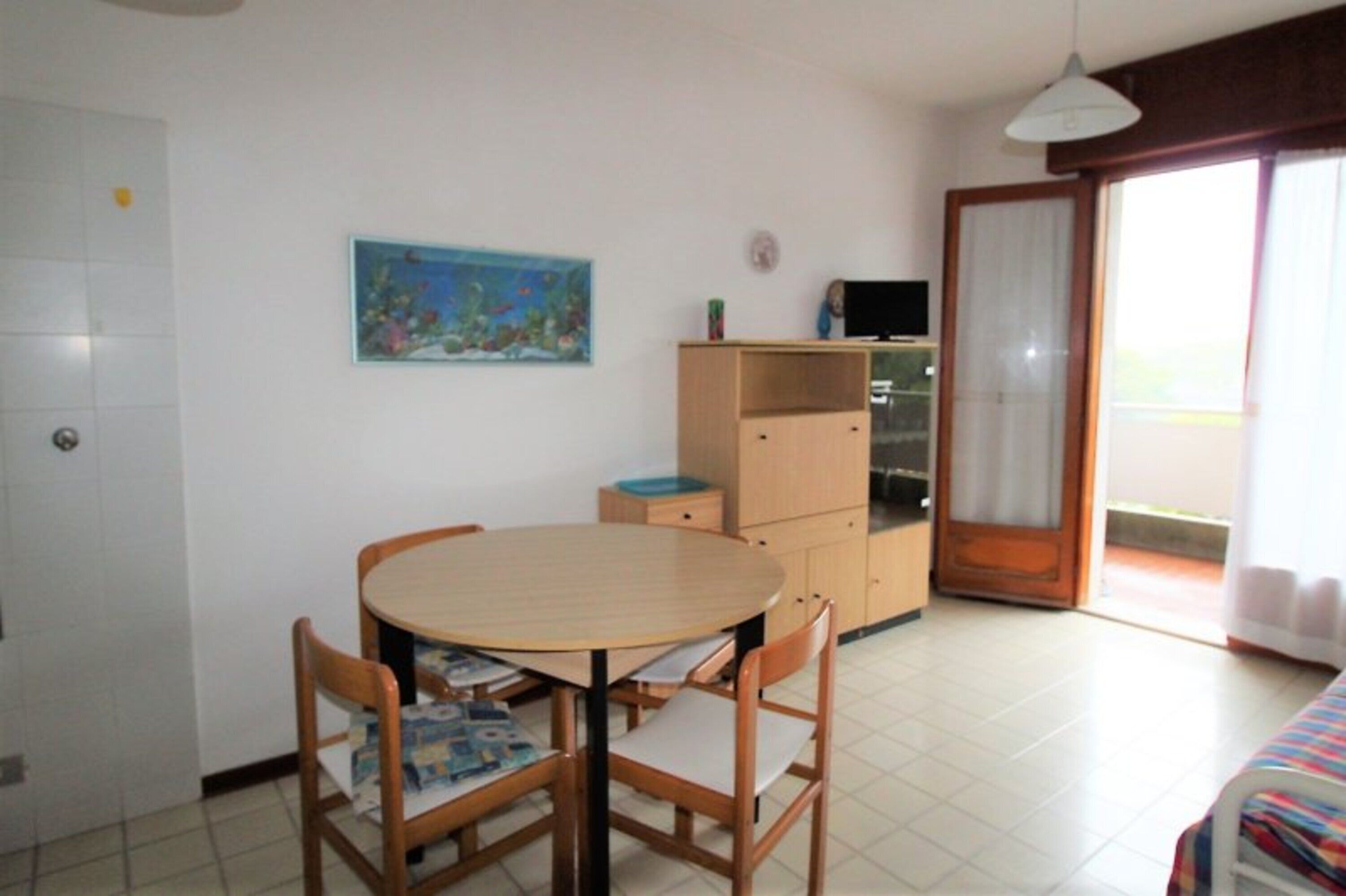 A room with a round wooden table, two chairs, a television on a stand, a painting on the wall, and a sliding door leading to a balcony.